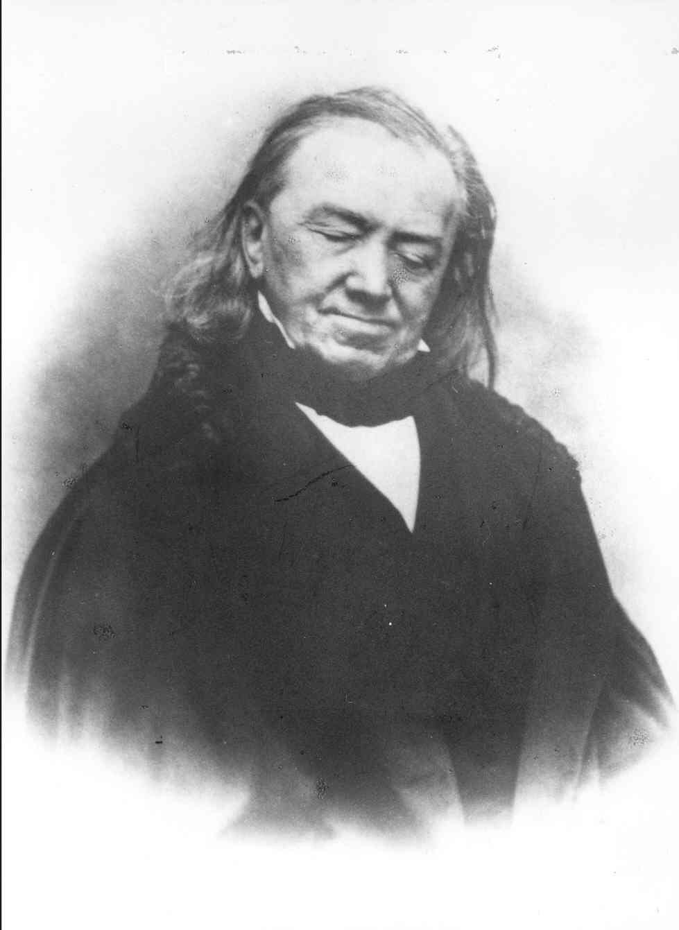 Porträt eines älteren Herren mit langen Haaren und geschlossenen Augen. Sein Gesicht ist zu einem leichten Lächeln verzogen und leicht zur rechten Seite geneigt. Er trägt dunklen Schal, weißes Hemd und schwarzen Mantel. Zu allen Seiten hin verliert sich die dunkle Fotografie im Weiß des Hintergrundes.